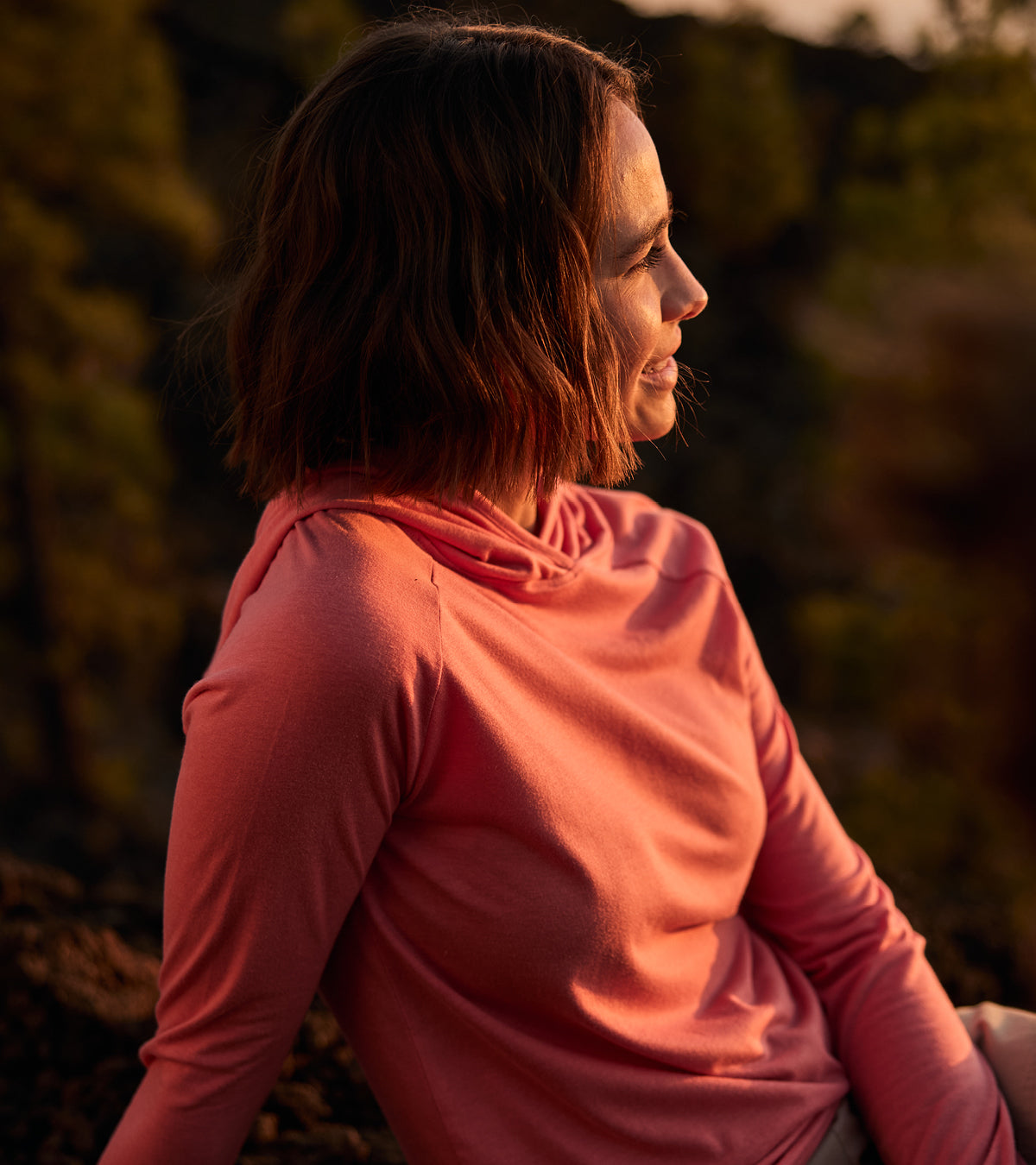 A woman in a pink lightweight hoodie sitting outdoors, relaxing on a rock with a blurred natural background. She is looking into the distance, with the orange light of the sun hitting her face.