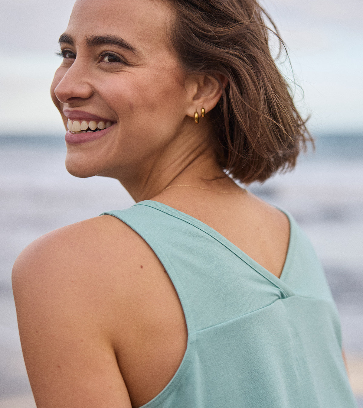 A close up of a woman with a light blue v-neck tank top standing by the ocean. She is looking over her shoulder, smiling at the camera.