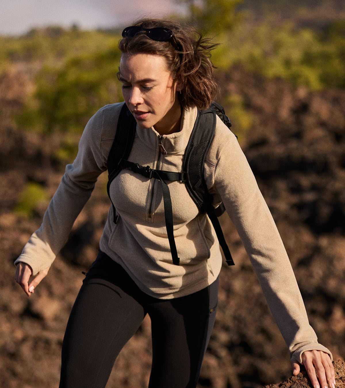 A woman person hiking outdoors with a backpack in a rocky landscape. She is wearing a pair of black leggings and a beige long sleeve fleece.