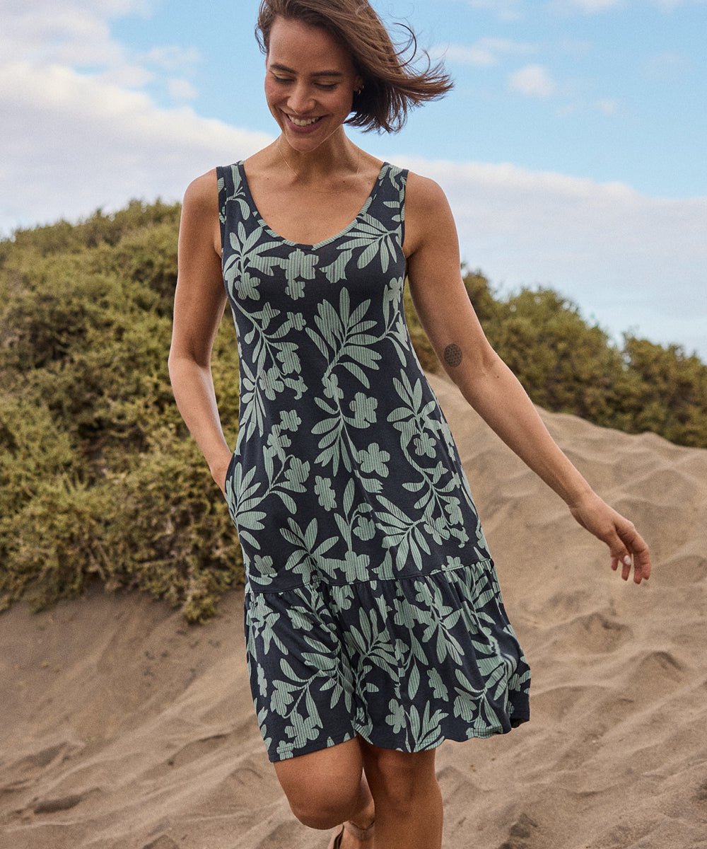 A woman in a black and green floral dress standing on a sandy beach with greenery in the background. She has one hand in her dress pocket and is smiling as she walks along the beach.