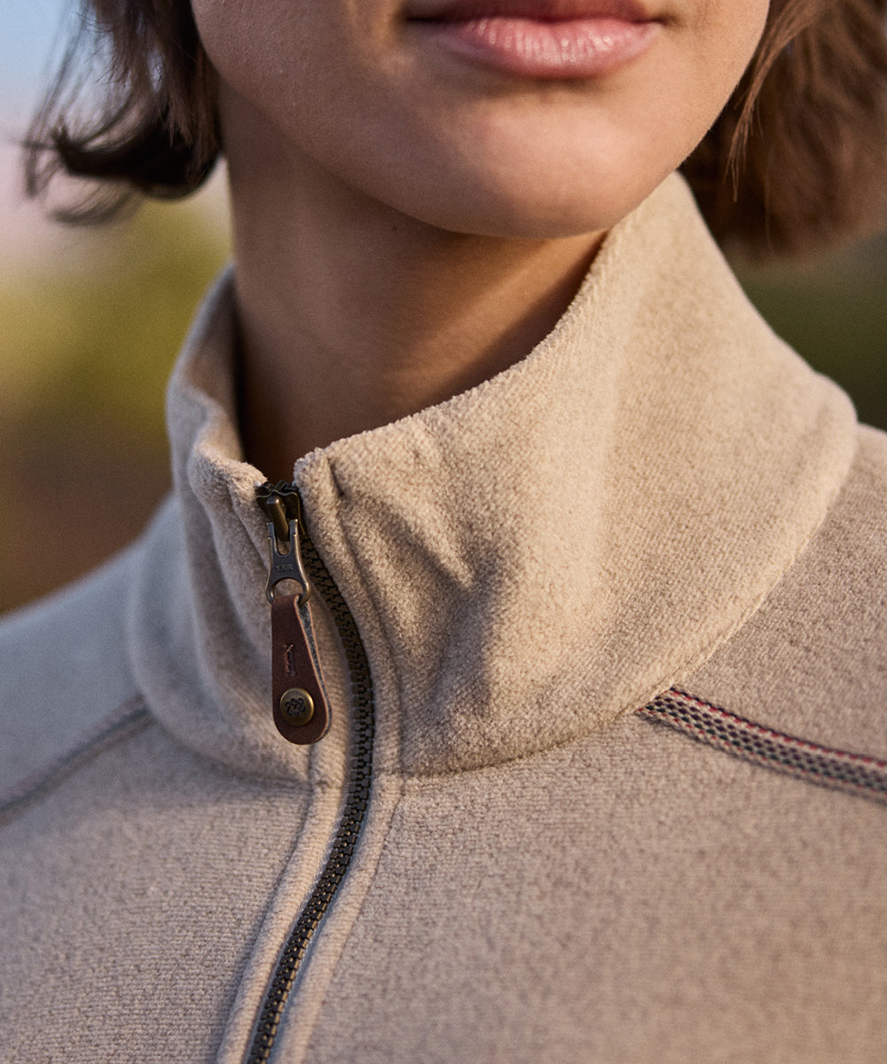 Close-up of a person wearing a beige fleece jacket with a blurred outdoor background. The fleece has a small leather puller with a metal rivet at the end.