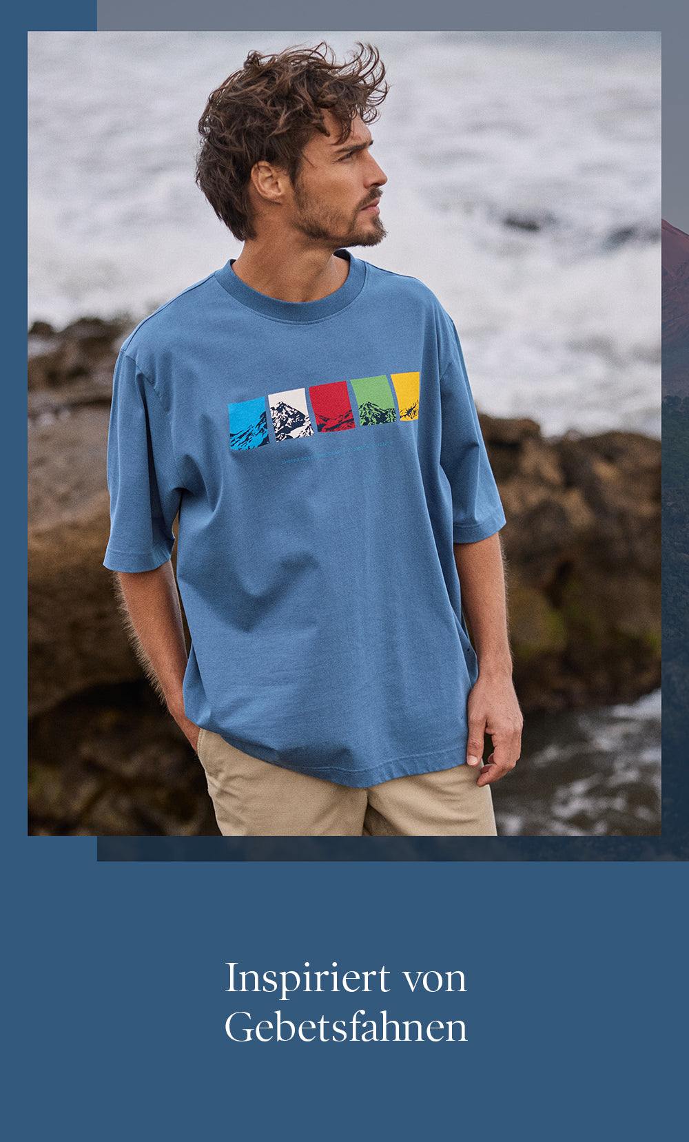 A wan wearing a blue t-shirt with colorful prayer flag and mountain design by a rocky coastline.