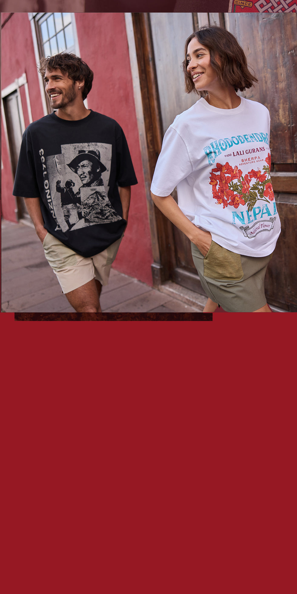 A man and a woman walking down a Spanish street. The man is wearing a black T-Shirt depicting Tenzing Norgay. The woman is wearing a white T-Shirt depicting a red rhododendron flower. They are smiling and have their hands in their pocket.