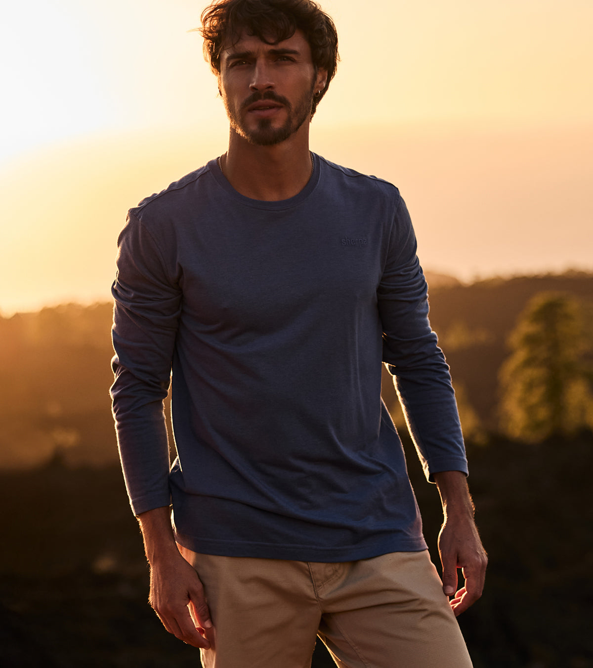 Man wearing a blue long-sleeve shirt and beige pants standing against a sunset landscape.