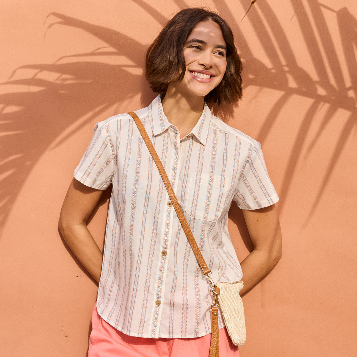 Woman wearing a striped shirt and pink pants against a peach-colored wall with palm leaf shadows.