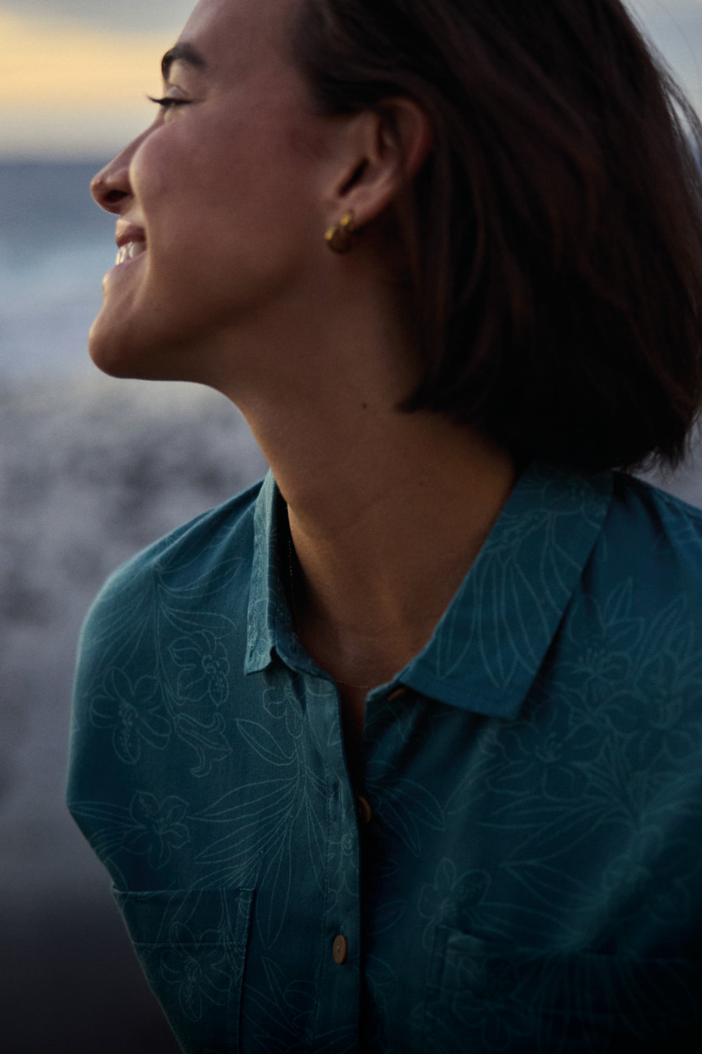 A close up of a woman in teal shirt standing on a beach at sunset. She is looking over her should smiling at the sunset.