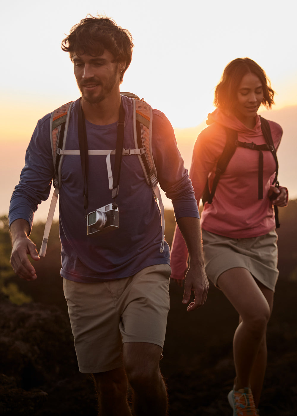 A man and a woman hiking on a hill at sunset. They have reached the crest of the hill and tree tops are visible around them. The man is wearing a blue long sleeve top, beige shorts, a backpack and a camera around his neck. The woman is slightly behind him, wearing a lightweight pink hoodie with a beige skort and a black backpack.
