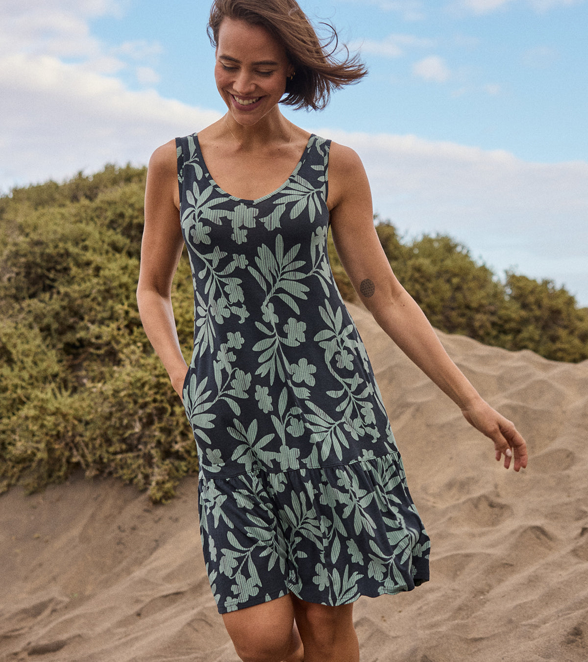 A woman walking on the beach sand dune in a black dress with a green floral pattern. She is smiling with one hand in her dress pocket.