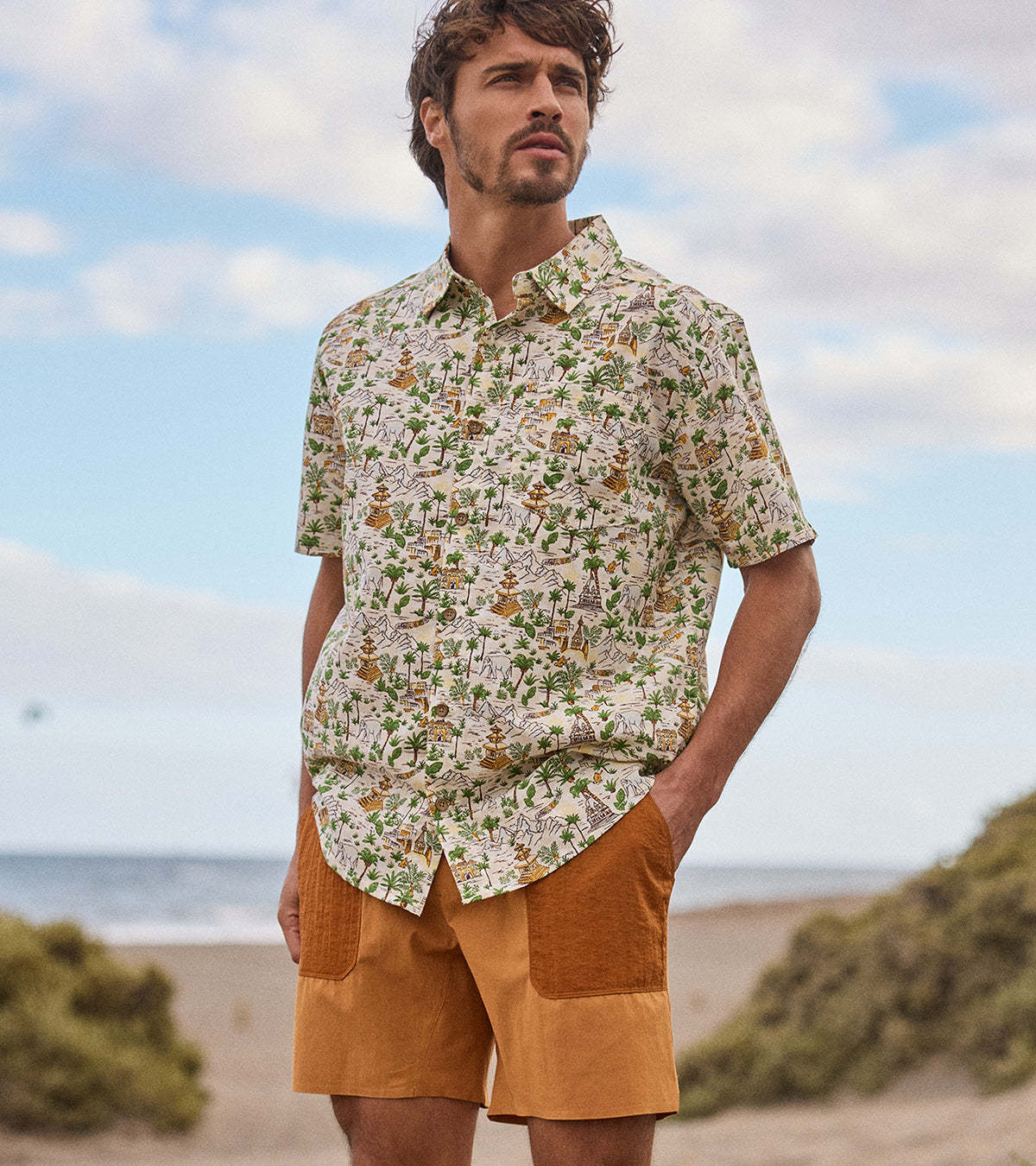 A Man wearing a patterned shirt depicting Nepal iconography such as temples and shrines. The shirt is paired with mustard yellow shorts with orange pocket overlays. He is standing on a beach on a warm summers day.