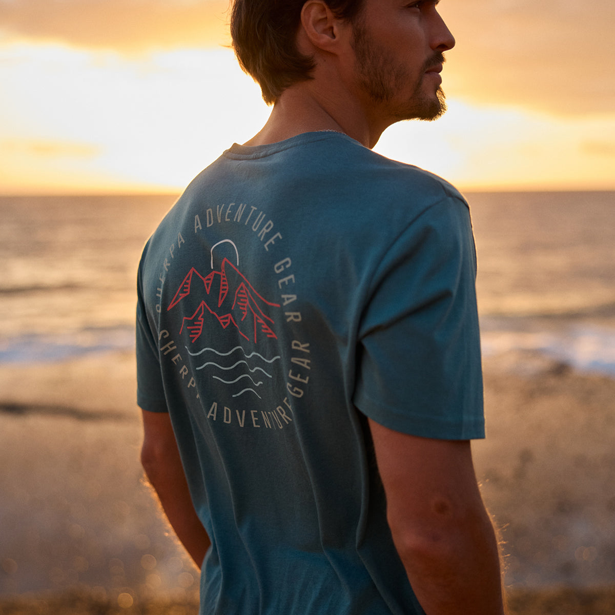 A mid shot of a man standing on a beach at sunset. He is wearing a teal blue t-shirt with a Himalayan mountain line graphic.