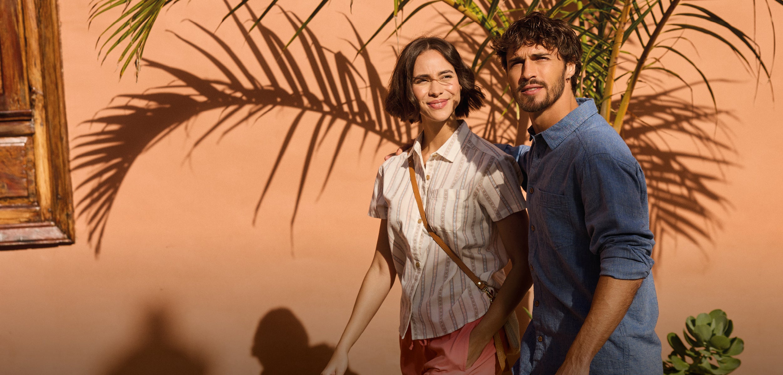 A man and a woman walking down a round in summer. The road has a peach coloured buildings and wall in the background with large potted plants. The man is wearing a lightweight blue shirt and a pair of long loose fitting trousers. The woman is wearing a white short sleeve shirt with vertical stripped pattern and pink shorts.