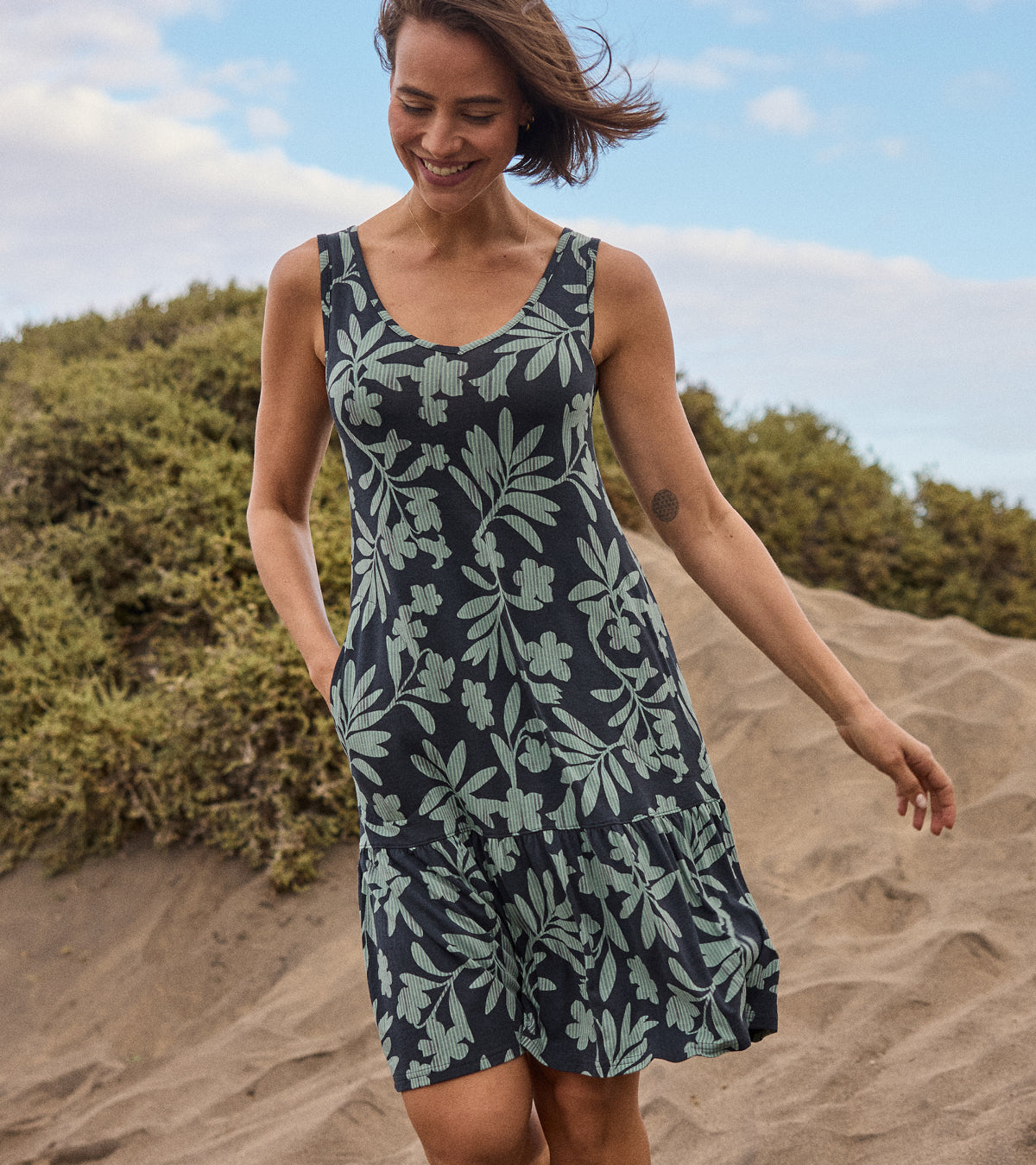 Woman wearing a floral dress on a beach with greenery and blue sky in the background