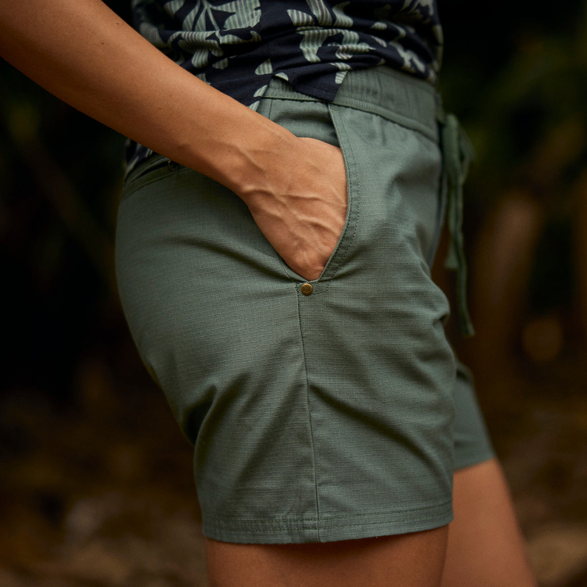 A close up of a woman wearing green shorts with a blurred natural background. The shorts are paired a black T-Shirt with a green floral pattern.