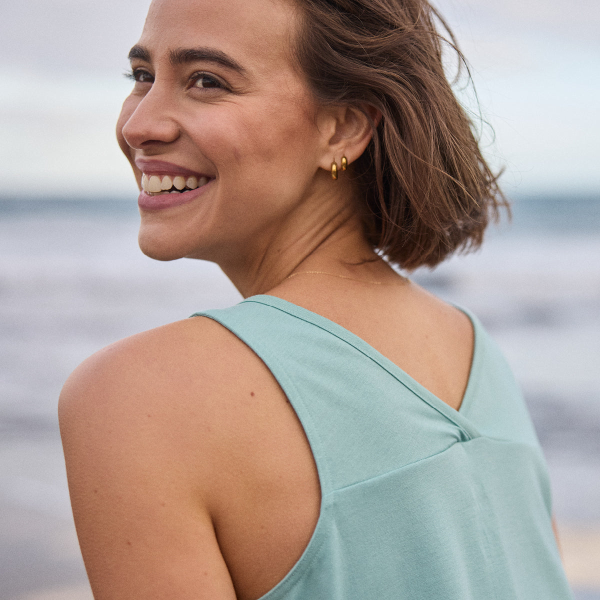 A close up of a woman in a light blue tanktop smiling with a blurred ocean background
