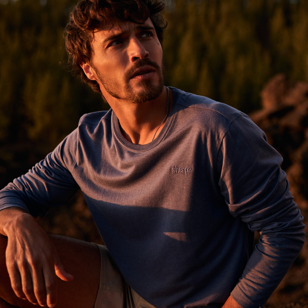 A man wearing a blue long sleeve shirt with an embroidered long logo, sitting outdoors with a blurred natural background. The golden light of a sunset is falling on him as he looks into the distance.