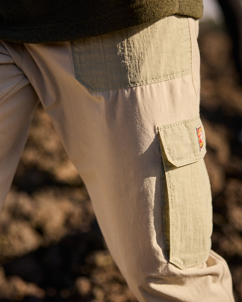 A close up of a man wearing a pair of beige cargo pants with the Sherpa Adventure logo on the pocket. The man is walking through a rocky landscape at sunset.