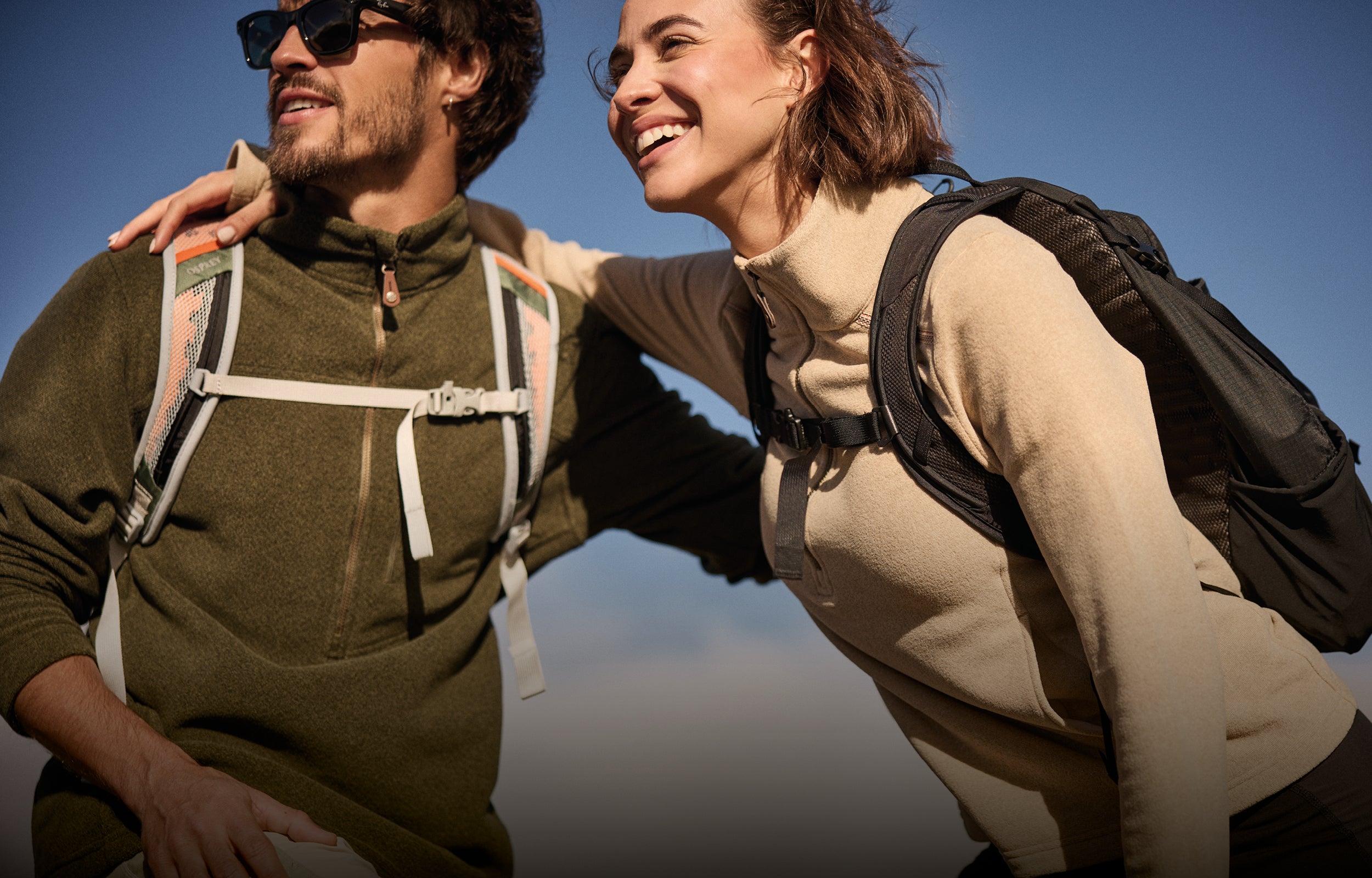 A man and a women with their arms around each other against a blue sky. They have been hiking and the man is wearing a green zip up fleece with beige hiking trousers. The woman is wearing a beige zip up fleece.