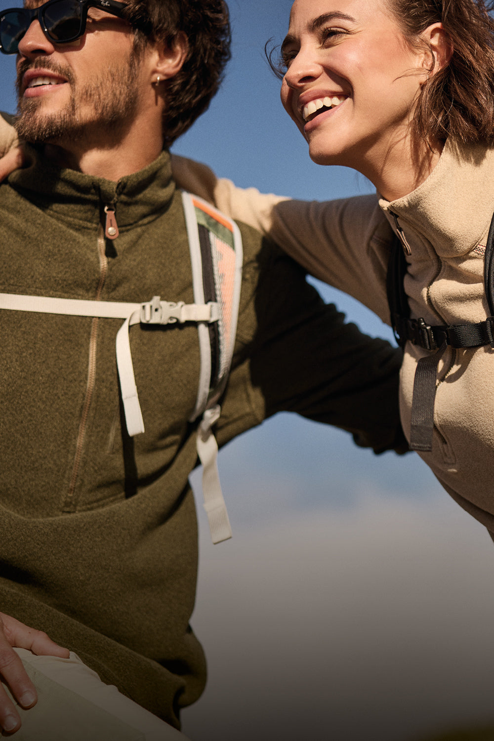 A man and a women with their arms around each other against a blue sky. They have been hiking and the man is wearing a green zip up fleece with beige hiking trousers. The woman is wearing a beige zip up fleece.