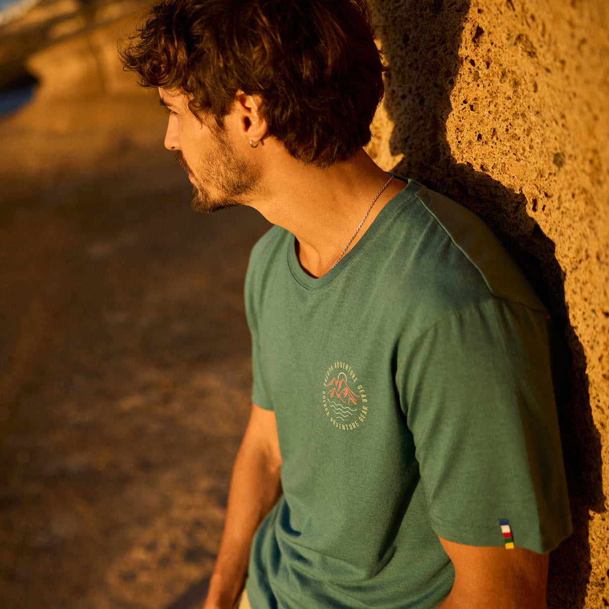 Man wearing a green t-shirt with a logo, standing against a textured wall. The logo depicts a red graphic mountain, a blue sea and white sun with the words "Sherpa Adventure Gear".