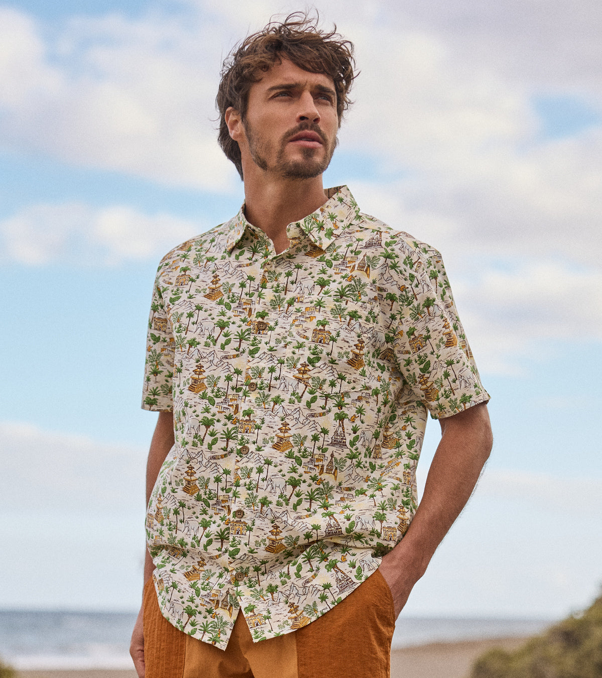 Man wearing a patterned shirt standing on a beach with a blue sky. The patterned shirt depicts palm trees and traditional Nepali temples.