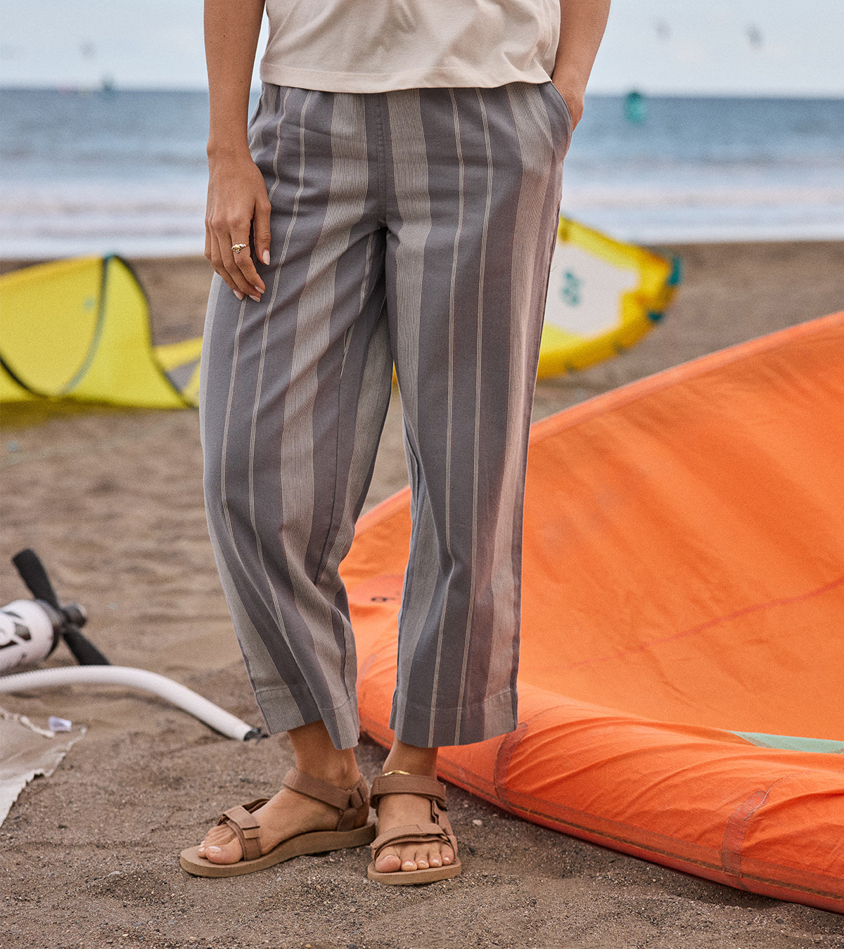 A woman wearing grey striped pants standing on a beach with orange and yellow kite in the background