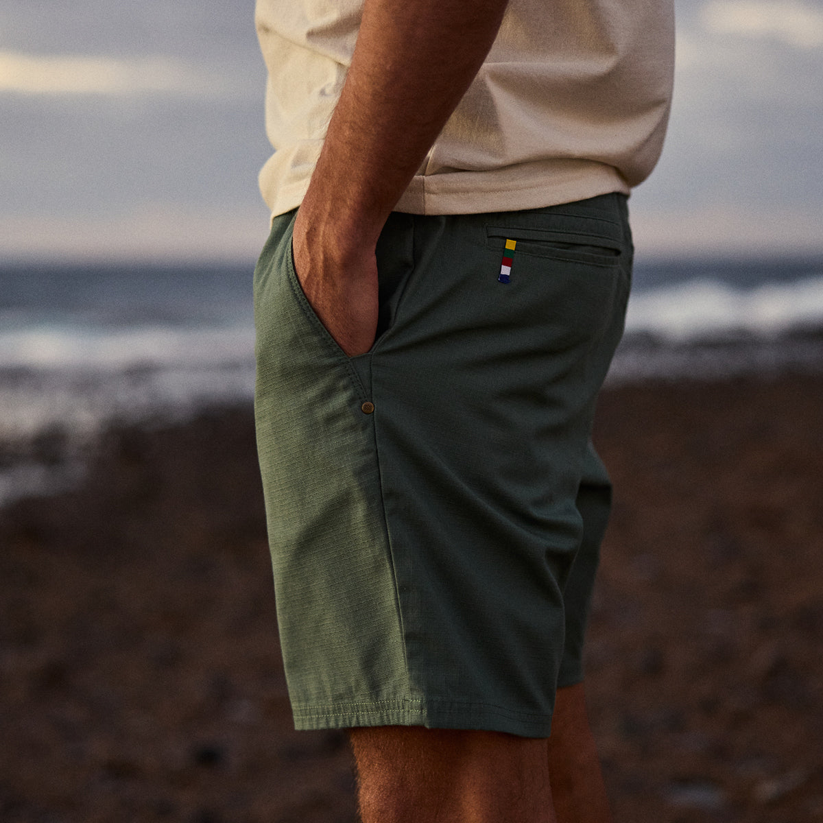 A man wearing green shorts with a blurred beach background. He is standing with his hands in his pockets.