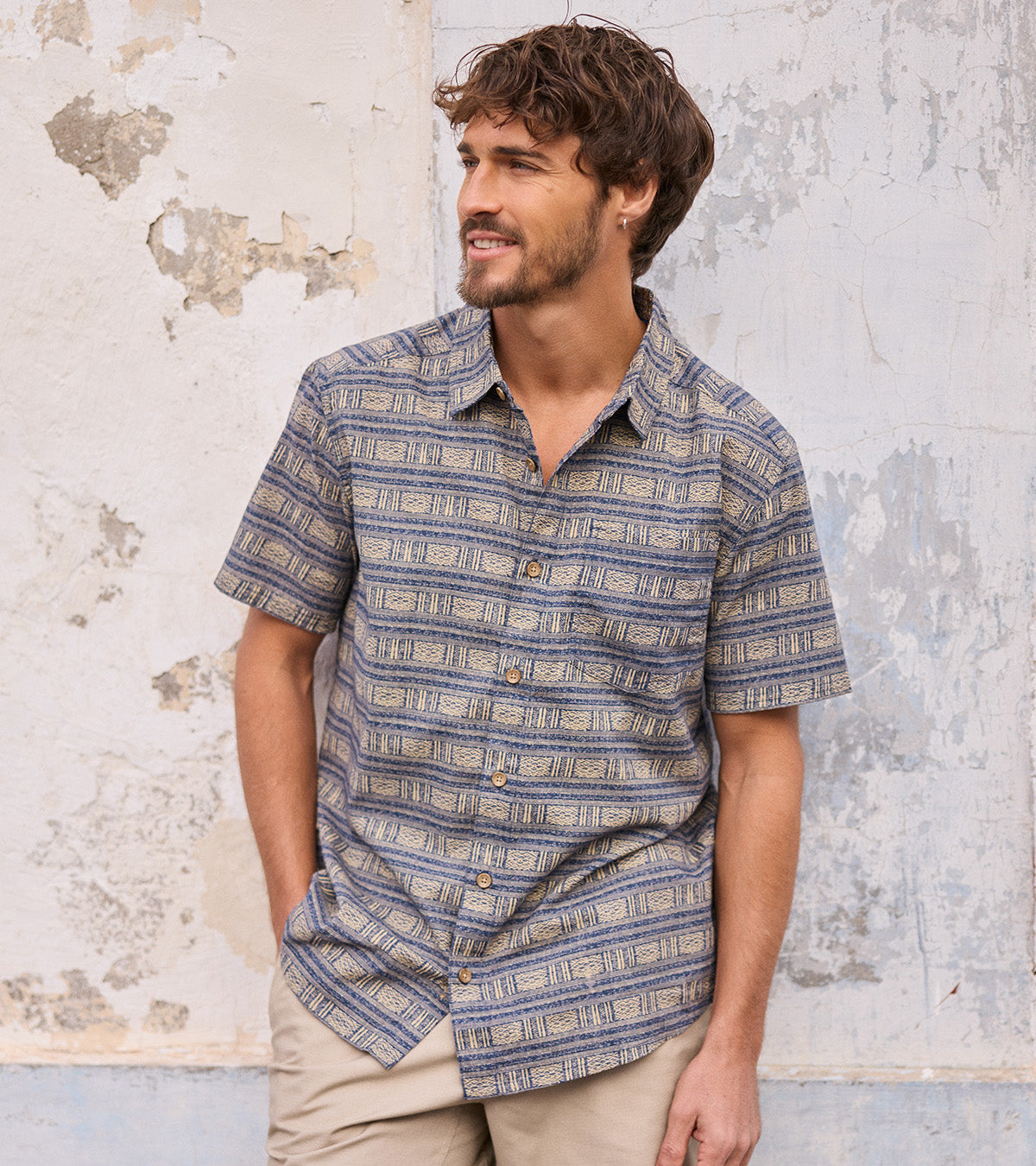 A man wearing a short-sleeve button-up shirt with a blue and beige geometric pattern, standing against a textured, peeling plaster wall.