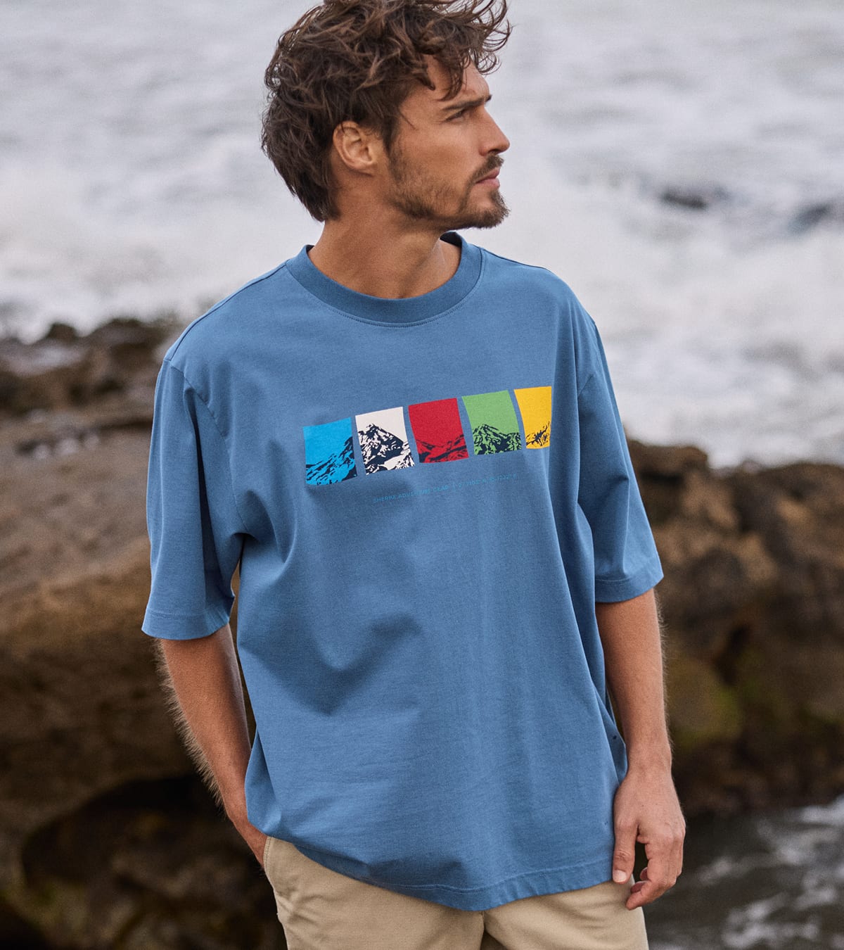 A man wearing a blue crewneck t-shirt with a colorful prayer flag graphic print across the chest, standing outdoors in front of a rocky, ocean background.