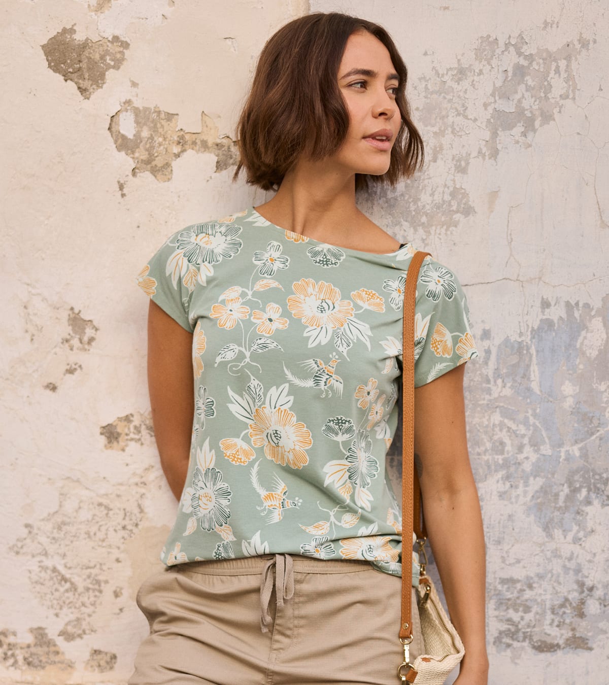 A woman wearing a sage green short-sleeve t-shirt with a white and yellow floral pattern, paired with beige drawstring pants and a cream shoulder bag, standing against a textured, peeling plaster wall.