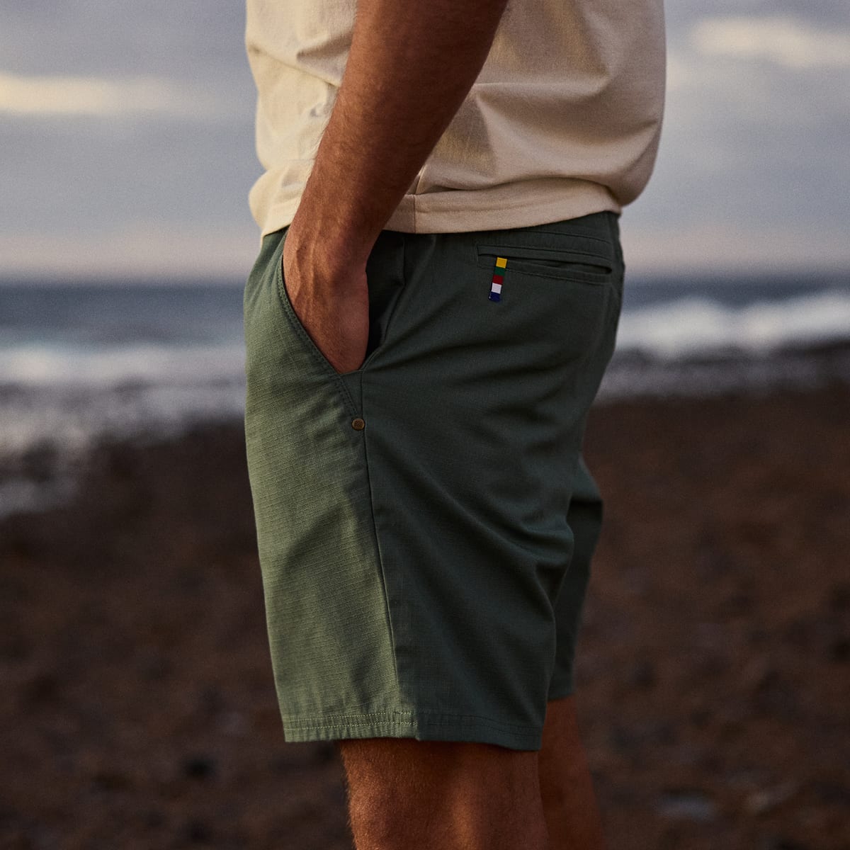 A man wearing dark green textured shorts and a light beige t-shirt standing on a rocky beach with the ocean in the background.