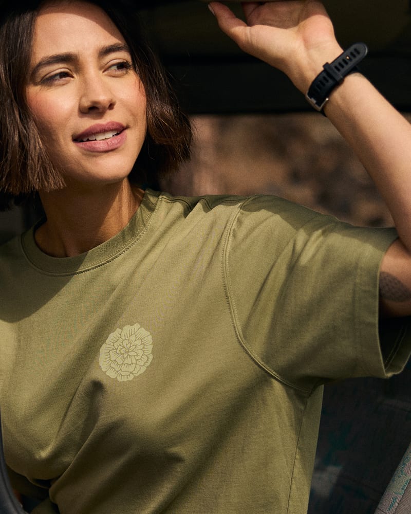 A woman wearing an olive green crewneck t-shirt with a light beige marigold floral graphic on the left chest, photographed in warm natural light.