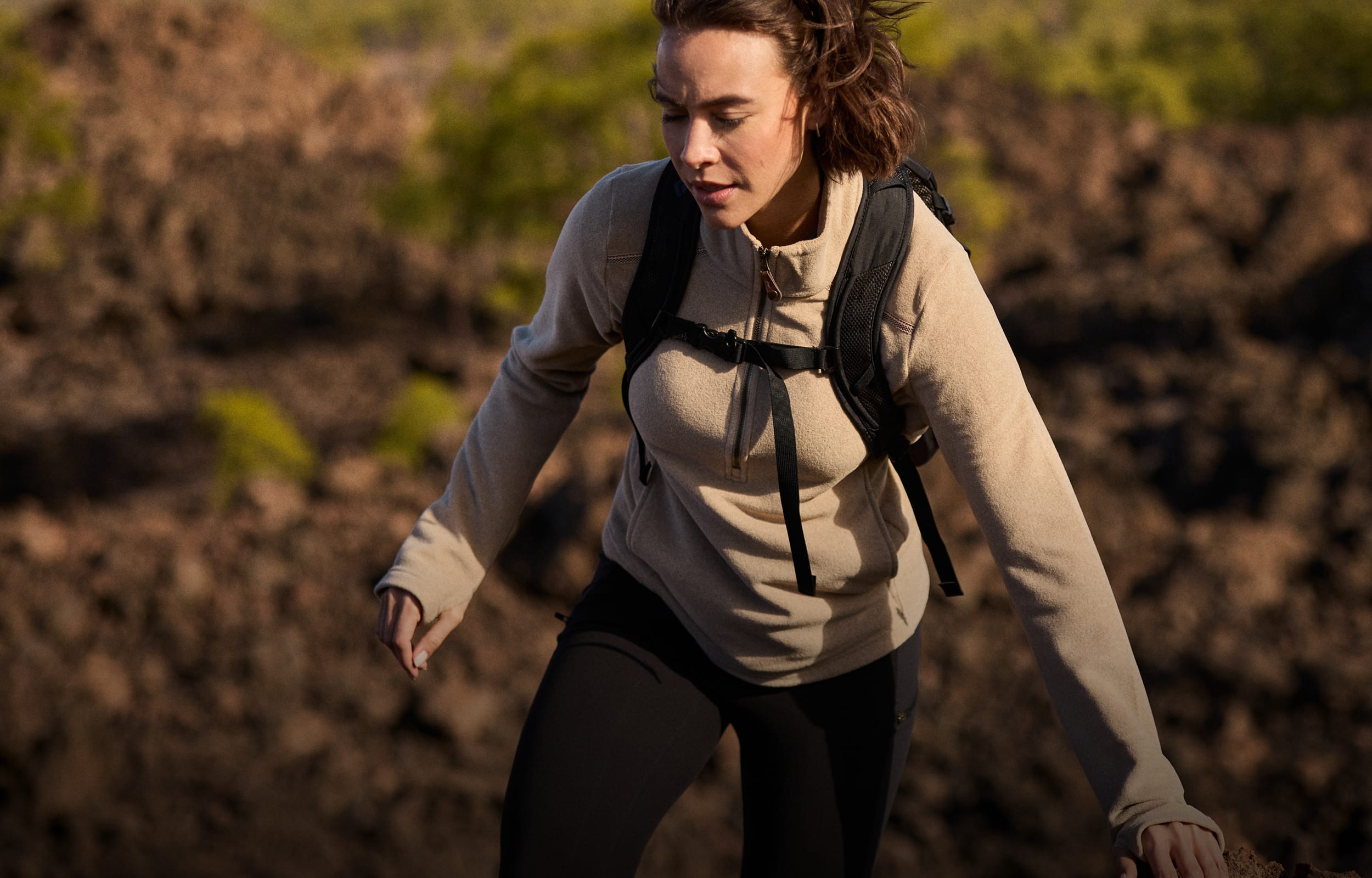 A woman hiking outdoors wearing a beige Sherpa Adventure Gear quarter-zip fleece, black leggings, and a black backpack, captured in warm natural light against a blurred brown and green landscape.
