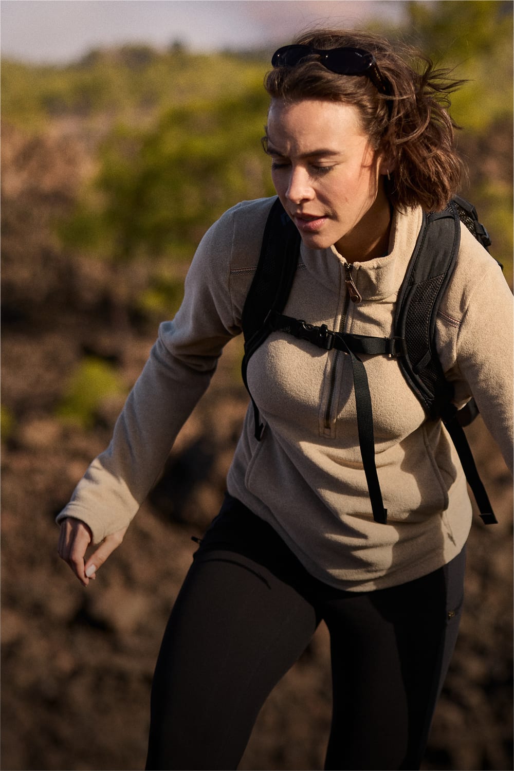 A woman hiking outdoors wearing a beige fleece pullover, black leggings, and a black backpack, with sunglasses on her head.