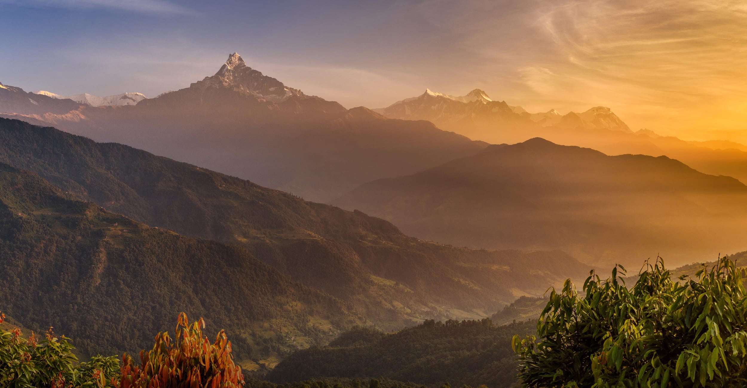 A scenic mountain landscape at sunrise or sunset, with layered green and brown ridges fading into mist, snow-capped peaks in the distance, and warm golden light across the sky.