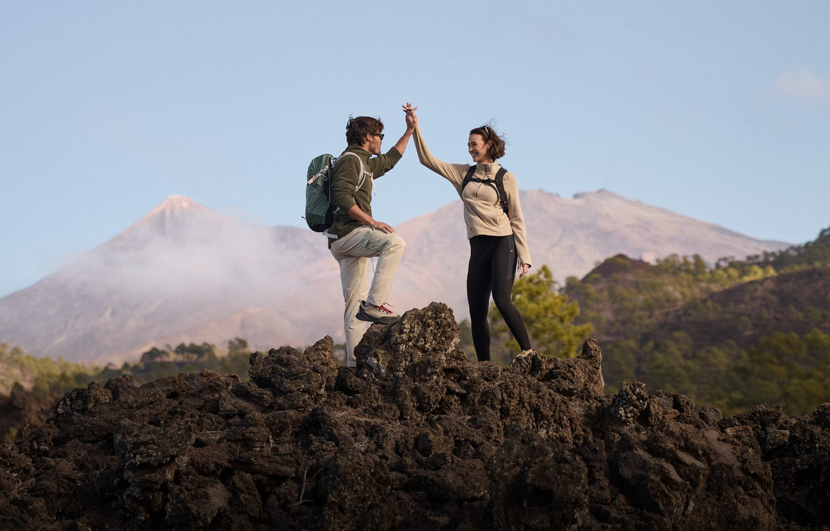 Two hikers high-five on a dark volcanic rock with mountains in the background under a clear blue sky. The man is wearing a green fleece, beige trousers and a green backpack. The woman is wearing black leggings, a beige fleece and a black backpack