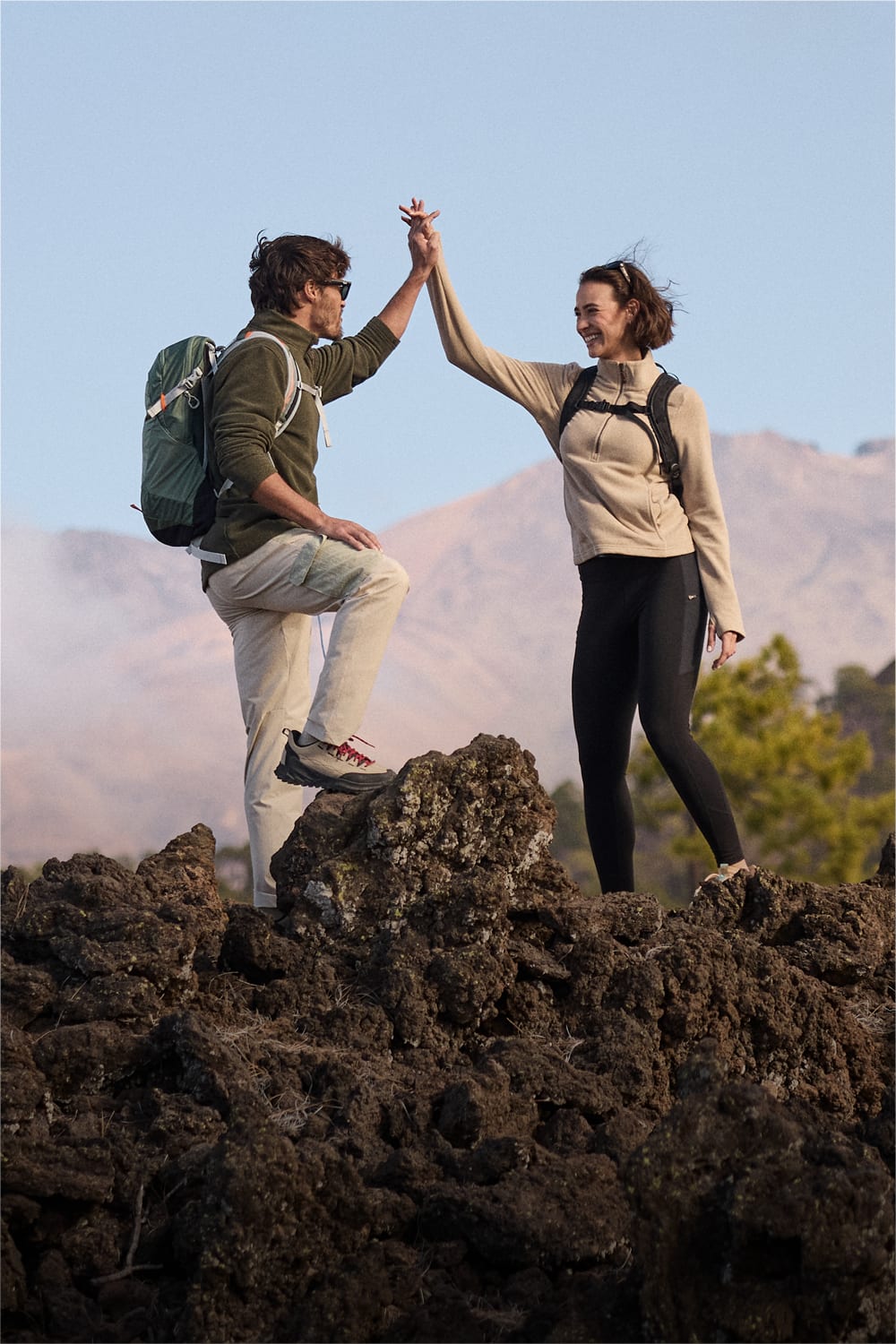 Two hikers high-five on a dark volcanic rock with mountains in the background under a clear blue sky. The man is wearing a green fleece, beige trousers and a green backpack. The woman is wearing black leggings, a beige fleece and a black backpack