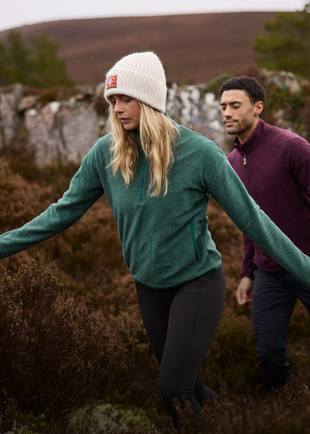 Woman in a white beanie and green quarter-zip fleece hiking outdoors, with a man in maroon behind her.