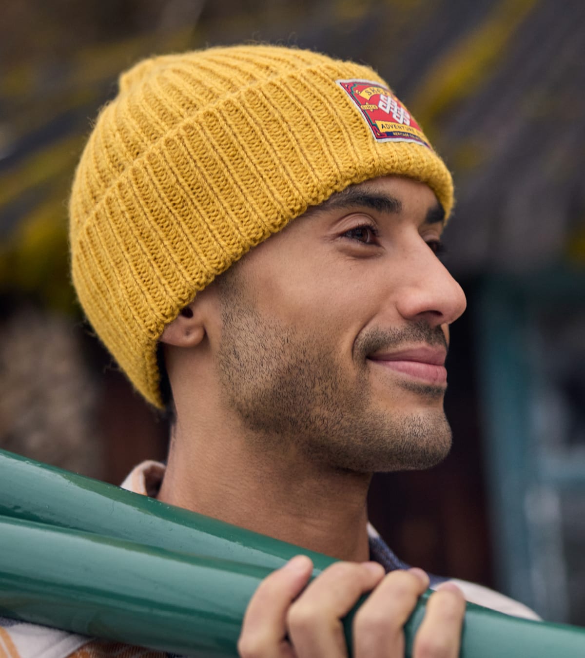 Man in a mustard yellow ribbed beanie with an embroidered patch, holding a green metal oar.