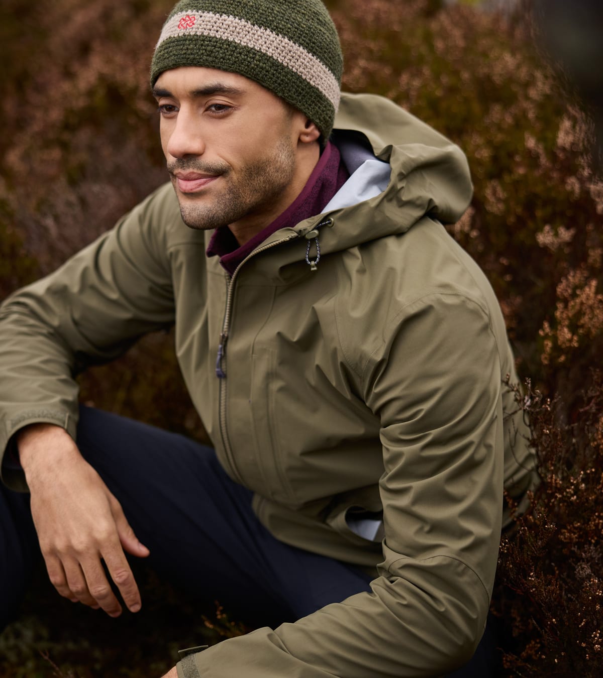 Man in an olive green hooded waterproof jacket and green beanie, crouching outdoors