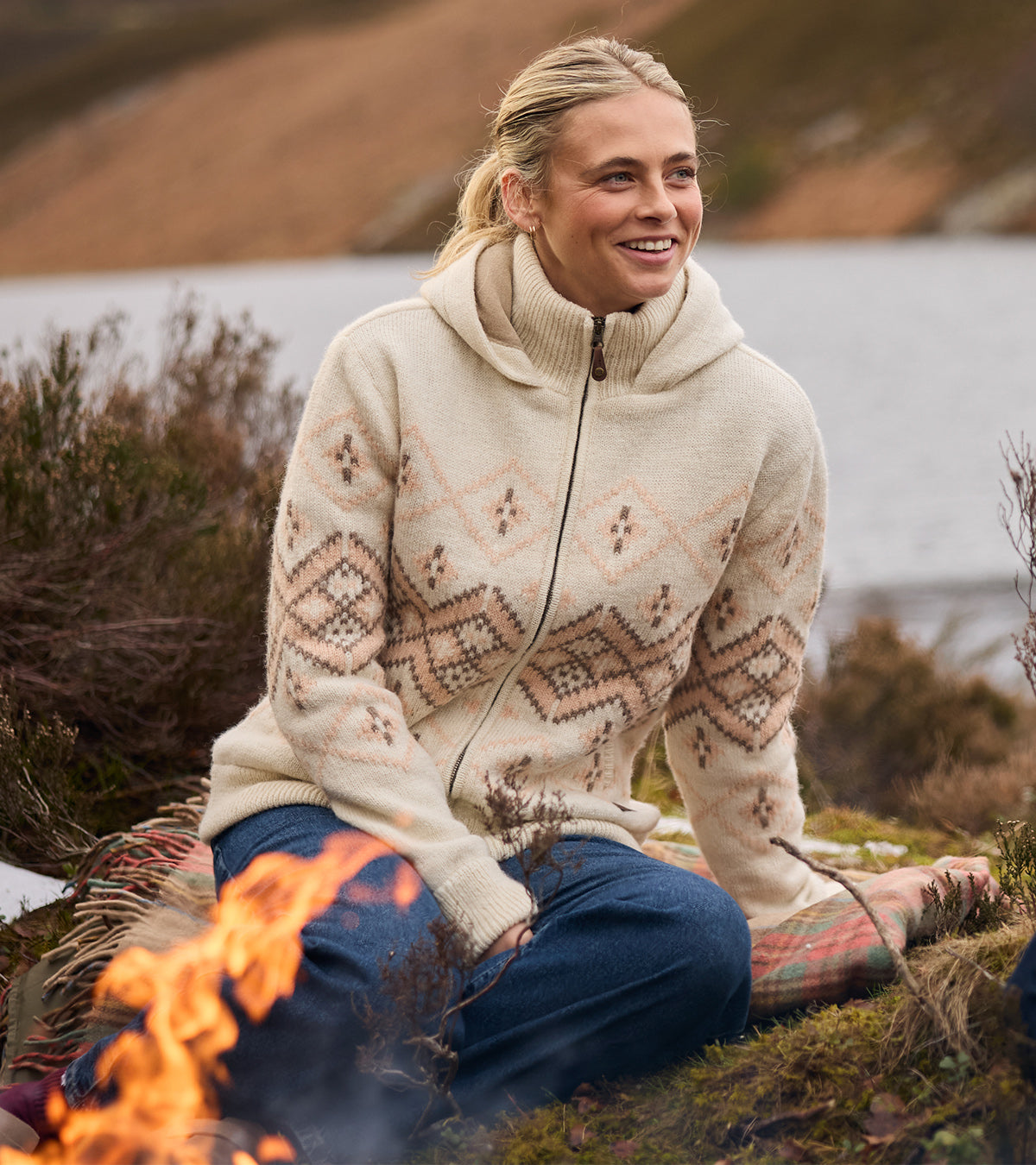 Woman sitting by a lake wearing a cream patterned knit hoodie and blue jeans.