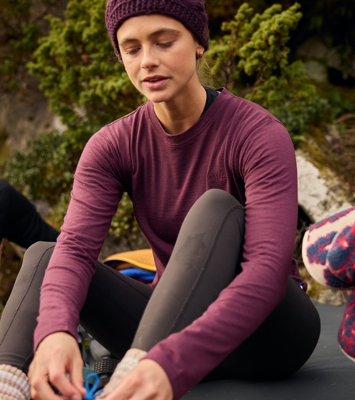 Woman in a maroon athletic top and knit beanie, sitting outdoors in greenery.