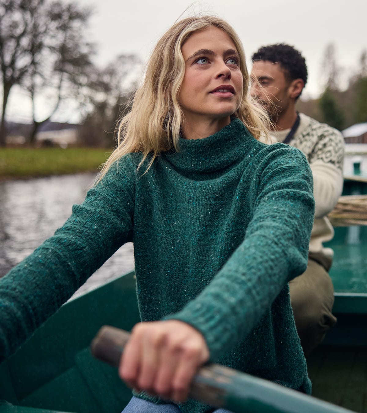 Woman in a green knit turtleneck rowing a boat, with a man in a patterned sweater behind her.