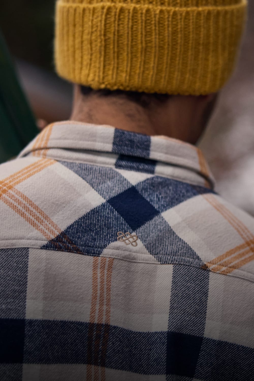 Person in a navy, cream and orange plaid flannel shirt with a yellow knit beanie, seen from behind.