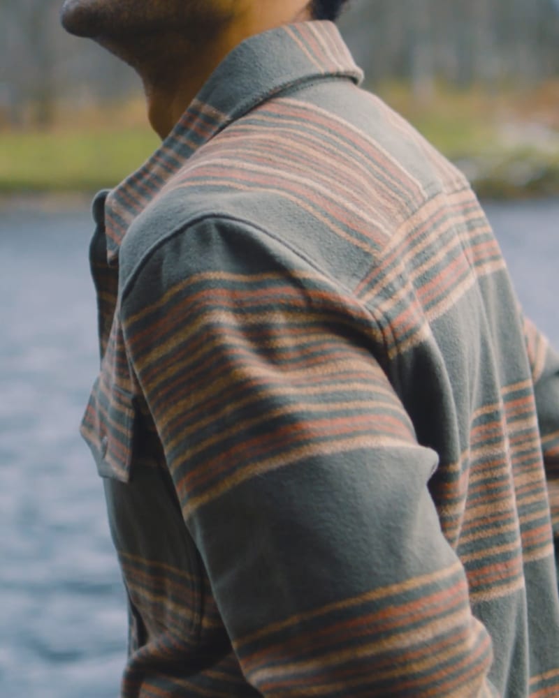 Man in a muted green striped flannel shirt with brown and orange accents, outdoors by water.