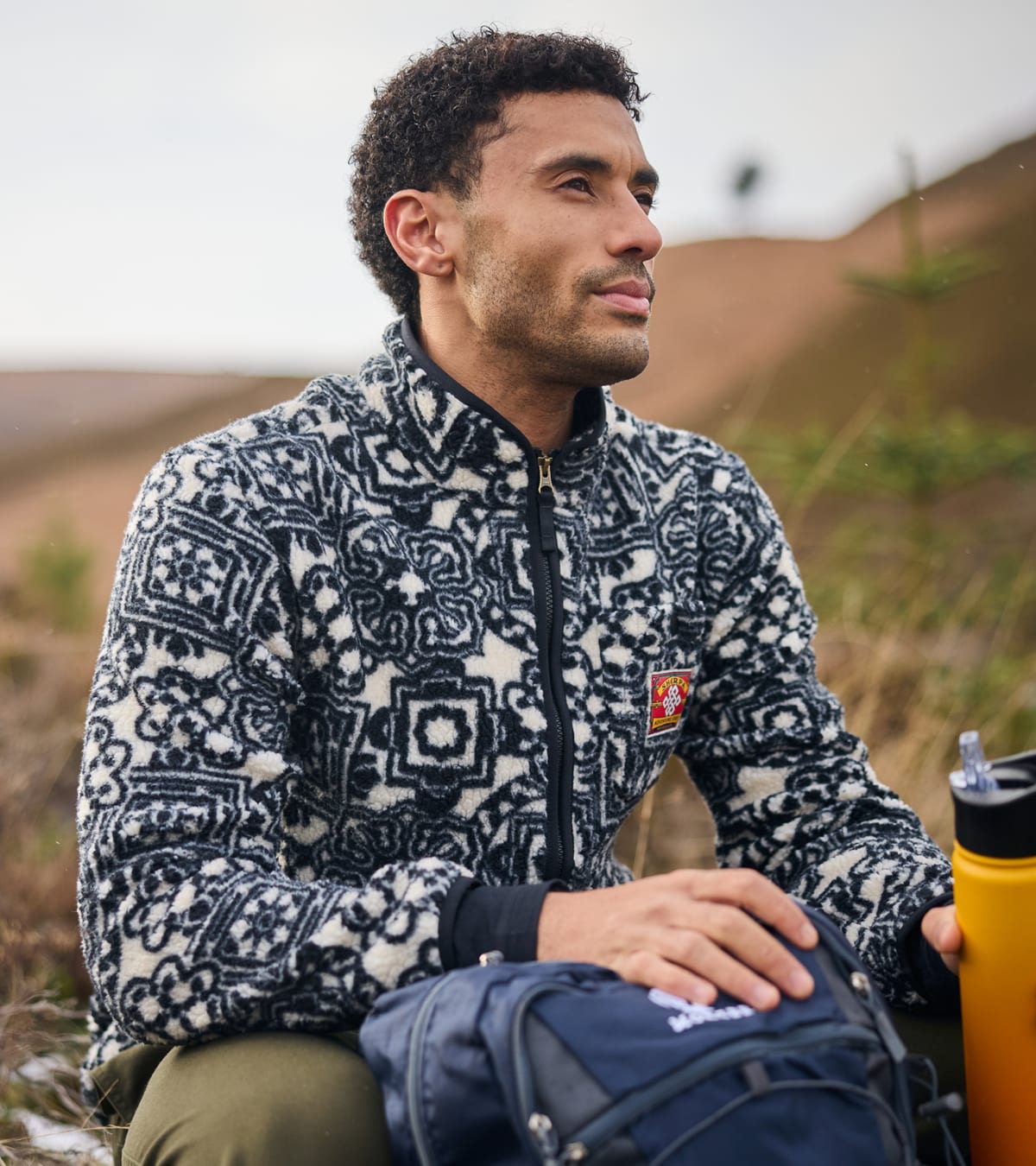 Man wearing a navy and cream patterned fleece, sitting on a grassy hillside with a backpack and water bottle.