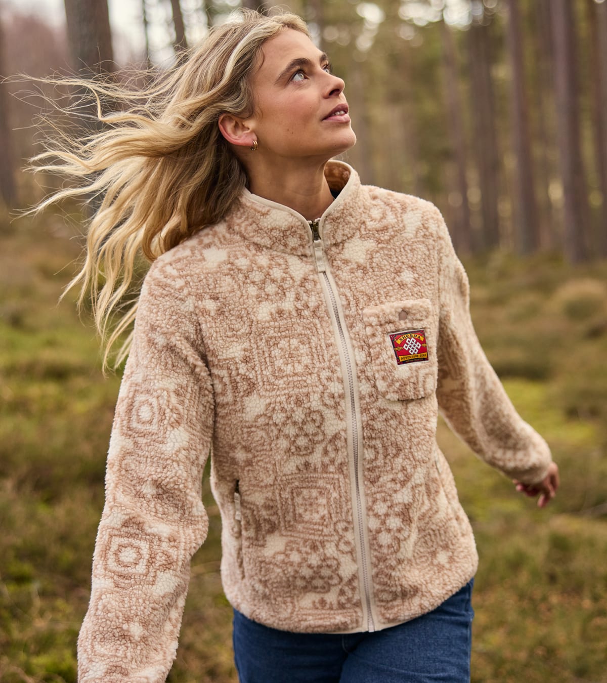 Woman wearing a beige and cream patterned fleece with a red and yellow logo patch, standing in a forest clearing.