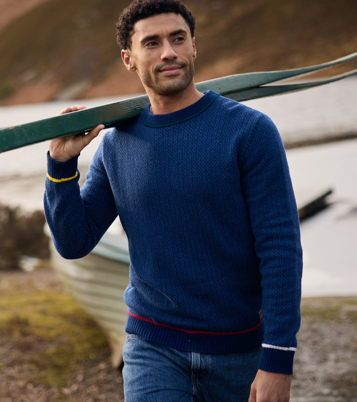 Man wearing a navy textured knit sweater with red and white trim, carrying a green boat outdoors.