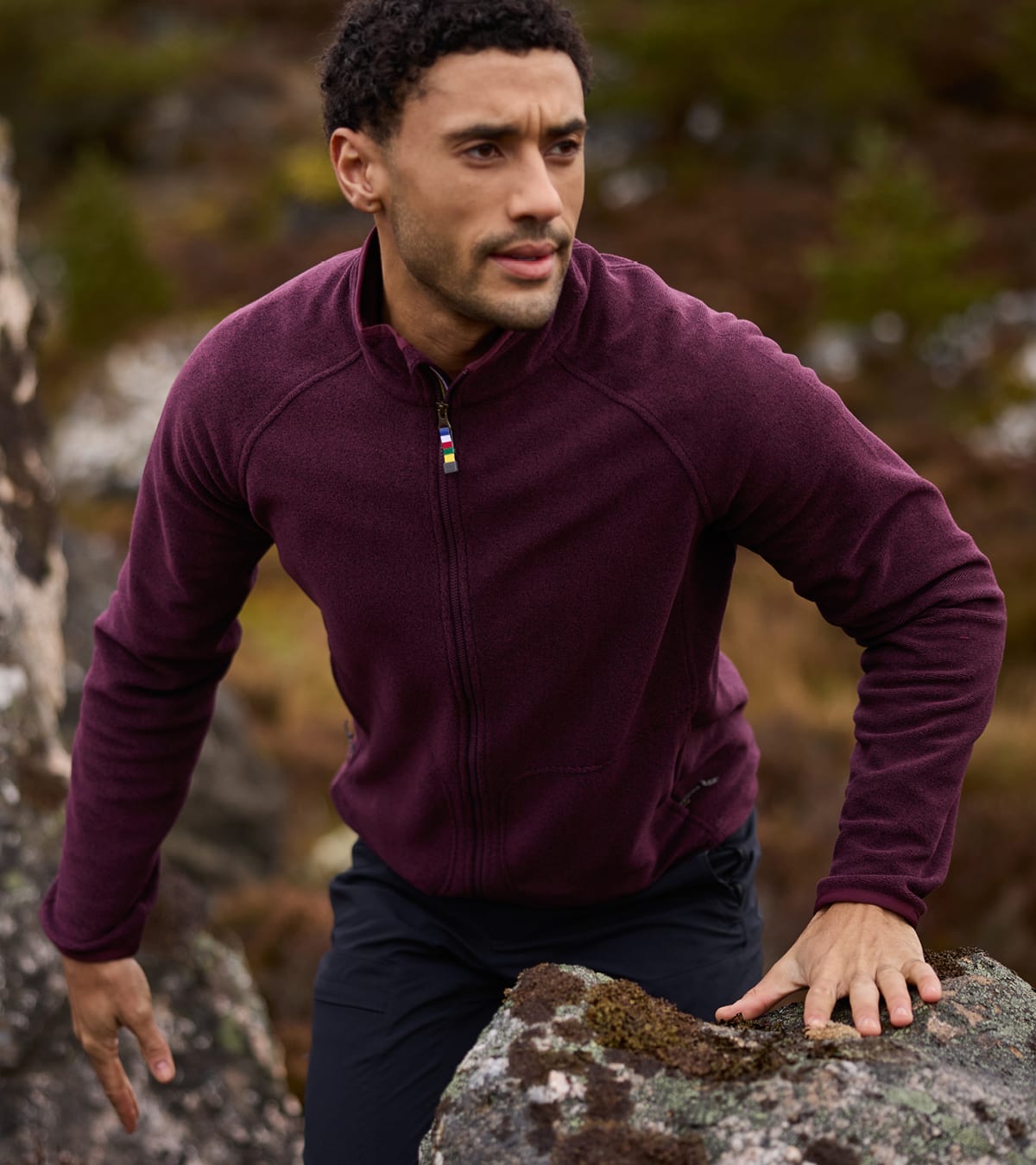 Man wearing a dark purple full-zip fleece and dark trousers, leaning on a mossy rock outdoors.