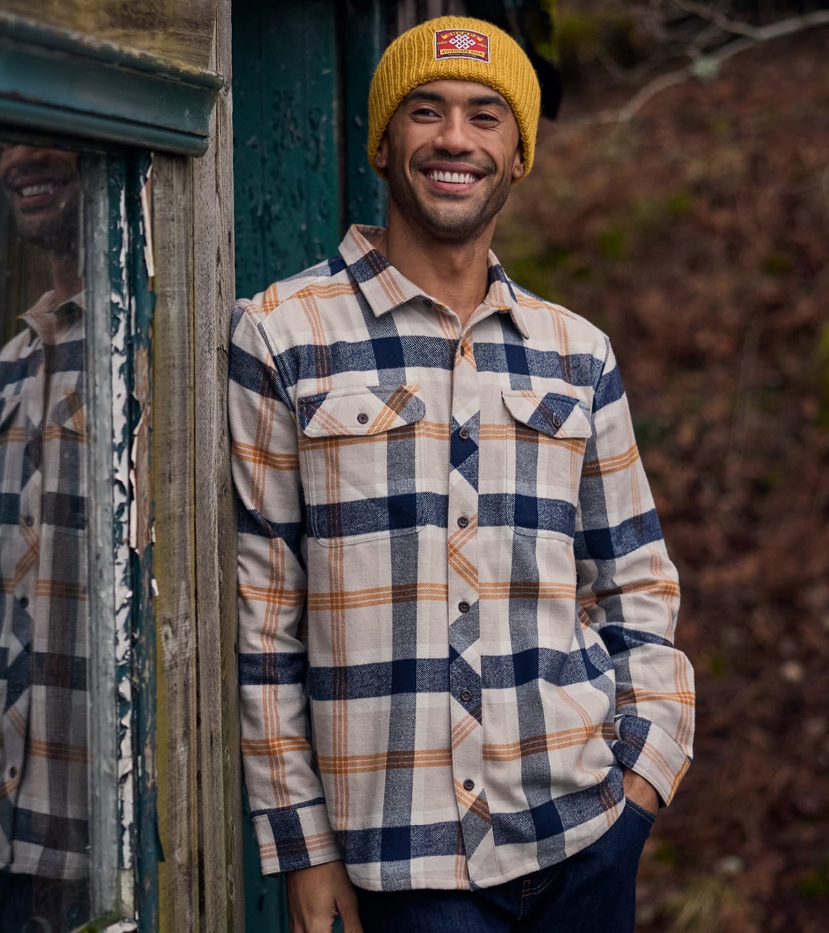 Man wearing a yellow beanie and navy, beige and orange plaid shirt, standing outdoors by a wooden door.