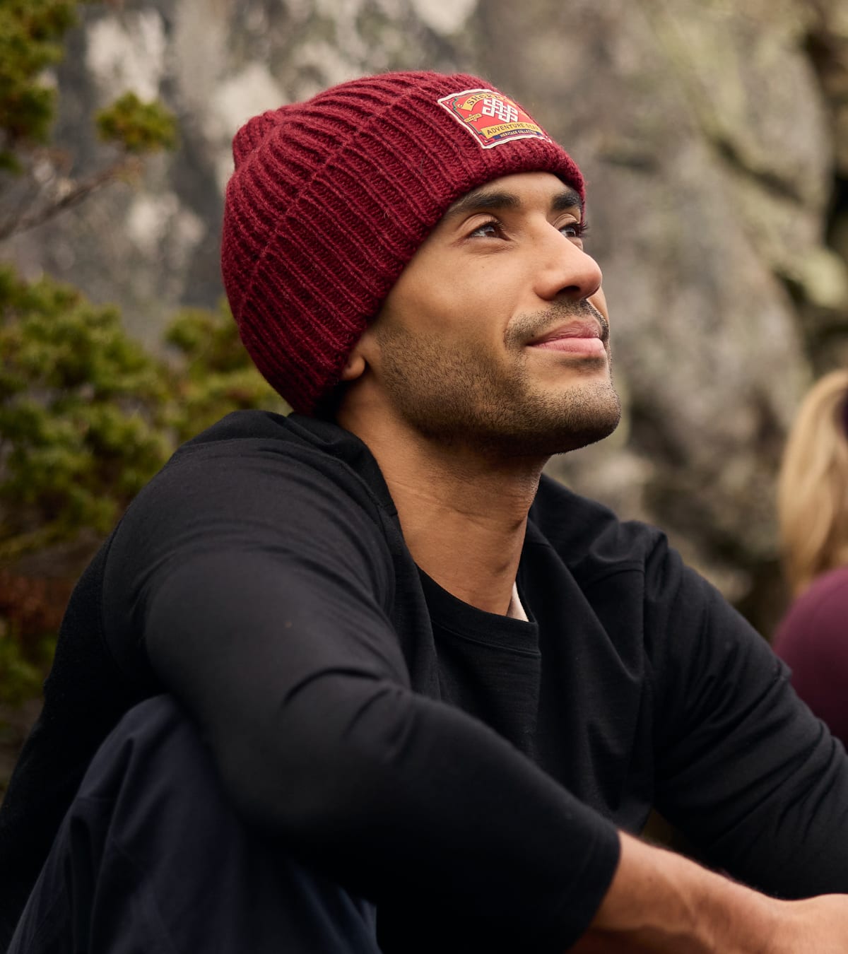 Man wearing a dark red ribbed knit beanie with a logo patch and black long-sleeve shirt outdoors.